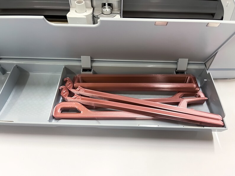 May include: A close-up of a gray drawer with a rose gold colored plastic tool inside. The tool is a long, thin, rectangular shape with a hook on one end.