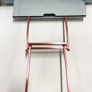 May include: A rose gold metal stand with two vertical supports and a horizontal bar. The stand is designed to hold a cutting mat.