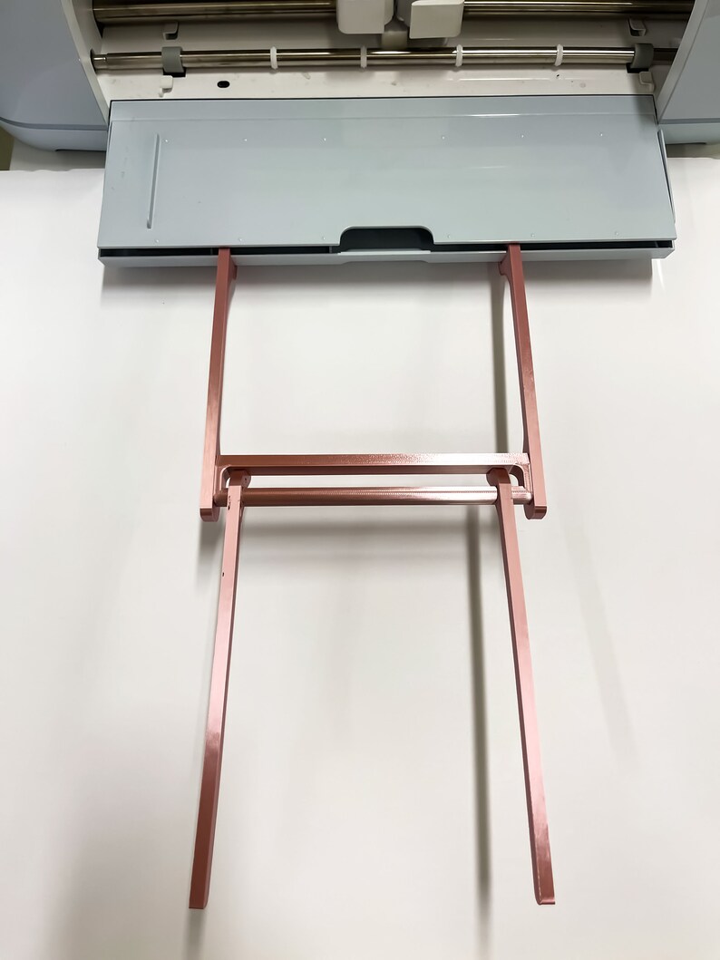 May include: A rose gold metal stand with two vertical supports and a horizontal bar. The stand is designed to hold a cutting mat.