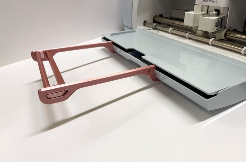 May include: A rose gold metal cutting mat holder for a Cricut Maker 3 machine. The holder is attached to the machine and has a handle on the side.