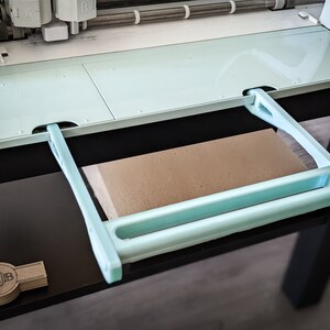May include: A light blue cutting machine with a retractable extension platform. The platform is made of plastic and has a light blue handle. The machine is sitting on a black surface.