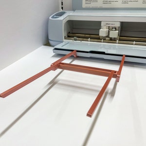 May include: A close-up of a light blue Cricut Maker 3 cutting machine with the cutting mat extended and the carriage raised. The machine is on a white surface. The carriage is made of metal and has a red, metal, folding stand.