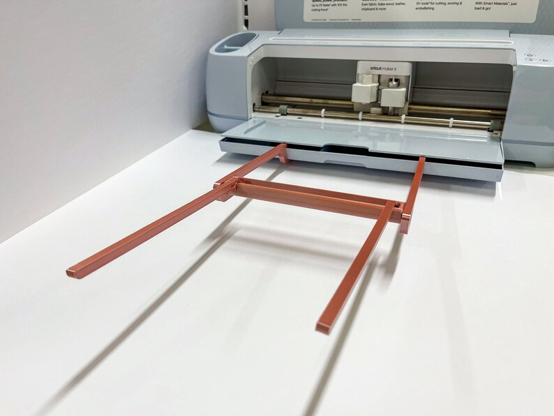 May include: A close-up of a light blue Cricut Maker 3 cutting machine with the cutting mat extended and the carriage raised. The machine is on a white surface. The carriage is made of metal and has a red, metal, folding stand.