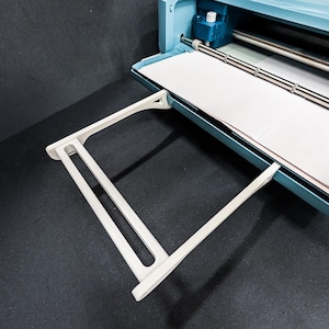 May include: A light blue cutting machine with a white paper sheet inside. A white support tray is extended from the machine. The machine has a silver roller and a blue control panel. The background is a dark gray surface.