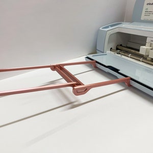 May include: A rose gold Cricut Maker 3 cutting machine with the cutting mat extended and the carriage in the home position. The machine is on a white surface.