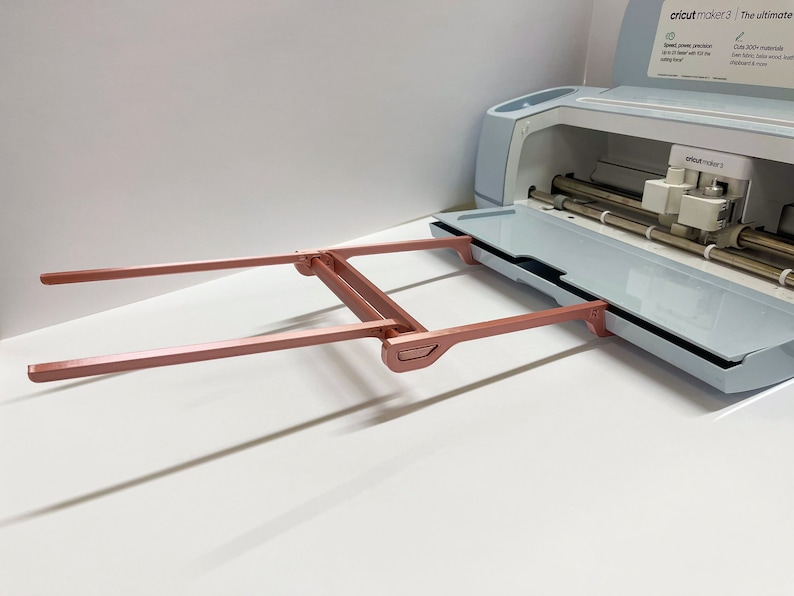 May include: A rose gold Cricut Maker 3 cutting machine with the cutting mat extended and the carriage in the home position. The machine is on a white surface.