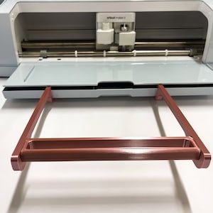 May include: A rose gold metal frame with a handle, designed to hold a cutting mat for a Cricut Maker 3 machine.