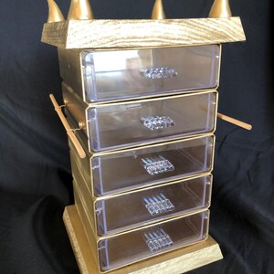 Op de afbeelding: Een goudkleurige decoratieve opbergunit met vijf doorzichtige lades, elk met een kristalachtige handgreep. De bovenkant heeft gouden hoorns en een gestructureerd oppervlak. De basis is ook goudkleurig. De tekst "Suggested Assembly" is zichtbaar.