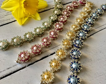 May include: Five beaded bracelets in different colors, including green, pink, yellow, and blue. Each bracelet features a central pearl bead surrounded by smaller beads in a flower-like design.