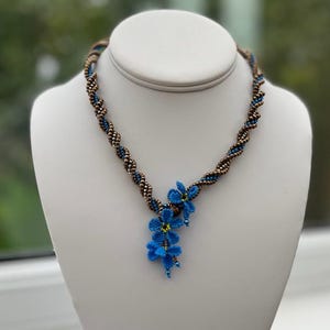 May include: A brown and blue beaded necklace with a blue flower pendant. The necklace is twisted and the flower pendant is made of small blue beads.