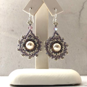Puede incluir: Un par de pendientes de plata con una perla y cuentas de color púrpura iridiscente. Los pendientes están colgados de un soporte blanco.