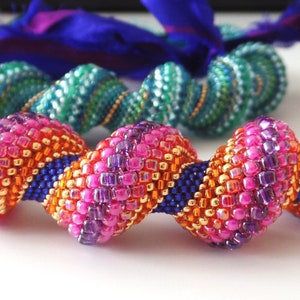 May include: A close-up of a beaded necklace with a spiral design. The necklace features vibrant pink, orange, and purple beads, accented with blue and gold. A second necklace with green and white beads is in the background.