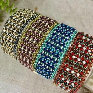 Puede incluir: Una colección de pulseras de cuentas en oro, morado, turquesa, azul y rojo. Cada pulsera presenta un patrón detallado y tejido con pequeñas cuentas. Las pulseras se exhiben sobre una superficie transparente. Circunferencia aproximada de 18 cm.