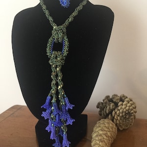 Könnte beinhalten: Eine lange grüne Perlenkette mit einem blauen Perlenakzent und einem Quasten aus violetten Perlen. Die Kette wird auf einer schwarzen Samt-Puppe präsentiert.