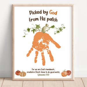 May include: A framed wall art print featuring the text Picked by God from His patch at the top, a central orange handprint shaped like a pumpkin with green vines, and the text For we are God's handiwork, created in Christ Jesus to do good works.