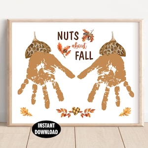 Op de afbeelding: Een ingelijste print met de tekst "Nuts about Fall" en twee handafdrukontwerpen. De handafdrukken zijn bruin, met illustraties van eikels en bladeren. Een zwarte ovaal onderaan leest "Instant Download".