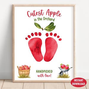 Op de afbeelding: Een ingelijste muurkunstprint met de tekst Cutest Apple in the Orchard bovenaan en Handpicked with love onderaan. Het midden toont twee rode aquarel babyvoetafdrukken onder groene bladeren, met een mand en een kruiwagen vol met appels.