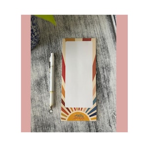 Puede incluir: Bloc de notas rectangular con un diseño retro que presenta un sol y las palabras "Happy Sunshine Days". El bloc de notas tiene un interior blanco y está bordeado por rayas verticales en tonos rojos, naranjas y azules. Un bolígrafo blanco descansa a su lado.