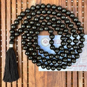 Puede incluir: Un largo collar de cuentas negras con una borla negra. Las cuentas son redondas y est&aacute;n espaciadas uniformemente. En el fondo se ve una peque&ntilde;a tarjeta con texto. Las cuentas probablemente se utilizan para la meditaci&oacute;n o las pr&aacute;cticas espirituales.