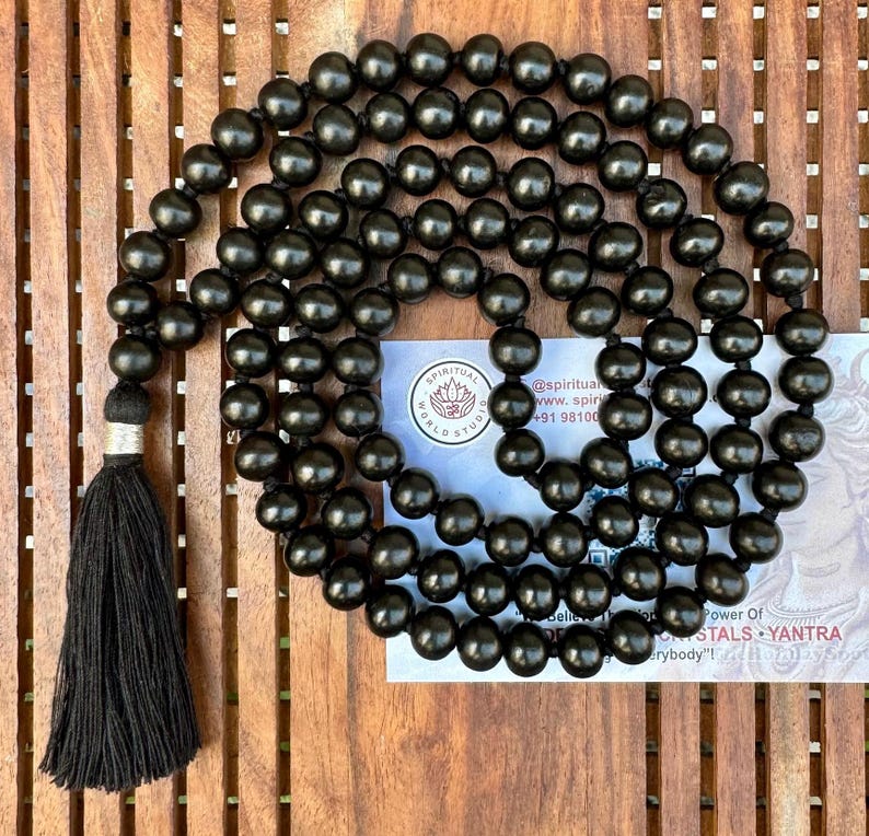 Puede incluir: Un largo collar de cuentas negras con una borla negra. Las cuentas son redondas y est&aacute;n espaciadas uniformemente. En el fondo se ve una peque&ntilde;a tarjeta con texto. Las cuentas probablemente se utilizan para la meditaci&oacute;n o las pr&aacute;cticas espirituales.