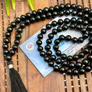 Puede incluir: Un collar de cuentas mala negro con una borla, sobre una superficie de madera. El collar presenta muchas cuentas redondas. Una tarjeta con texto es parcialmente visible, incluyendo las palabras "SPIRITUAL WORLD STUDIO".