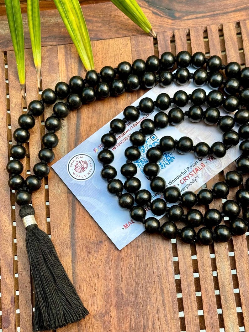 Puede incluir: Un collar de cuentas mala negro con una borla, sobre una superficie de madera. El collar presenta muchas cuentas redondas. Una tarjeta con texto es parcialmente visible, incluyendo las palabras "SPIRITUAL WORLD STUDIO".