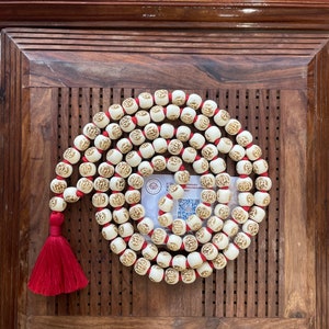 May include: A white mala bead necklace with red string and a red tassel. The beads are round and have a raised design. The necklace is on a wooden surface.