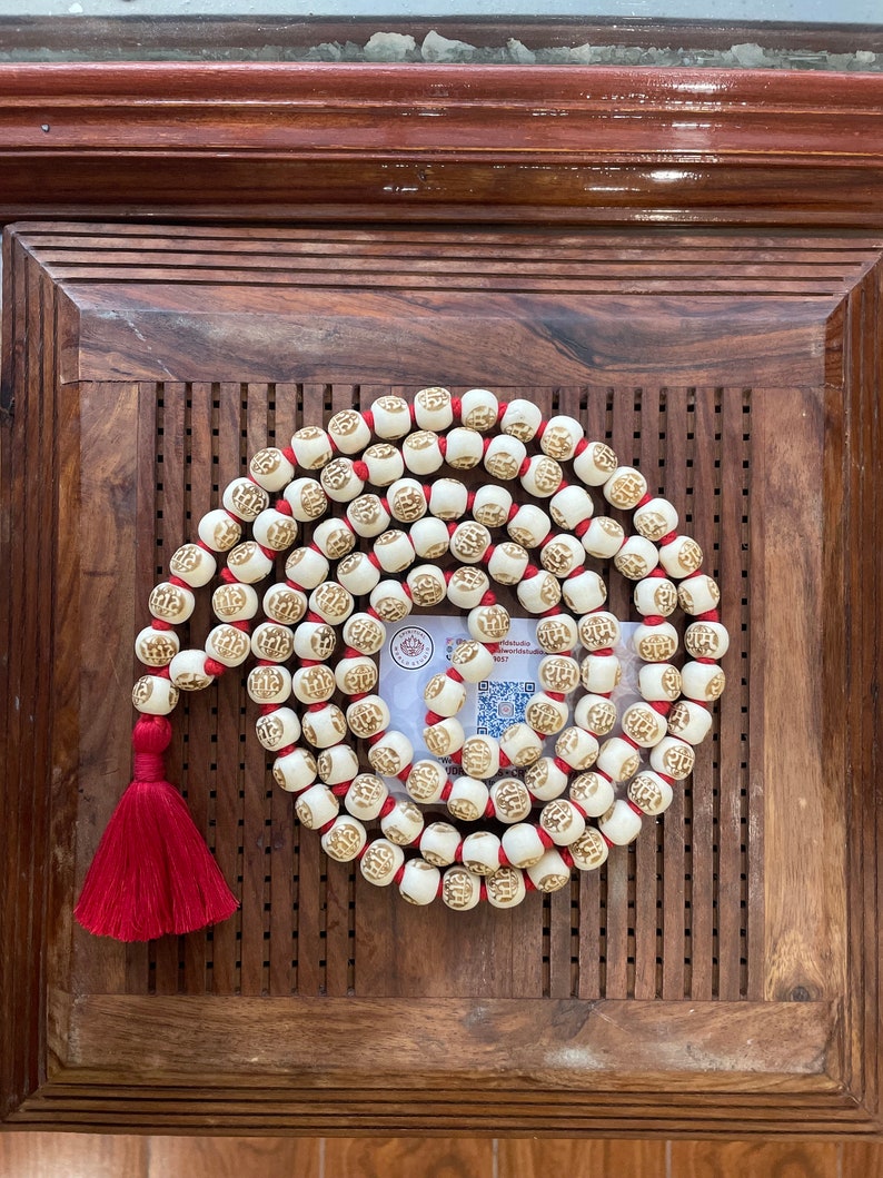 May include: A white mala bead necklace with red string and a red tassel. The beads are round and have a raised design. The necklace is on a wooden surface.
