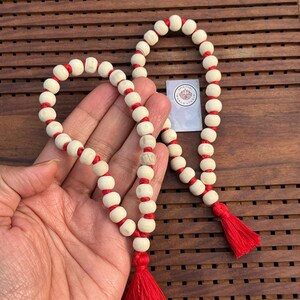 May include: A white and red prayer beads mala with a tassel on each end. The beads are made of wood and are strung together with red thread.