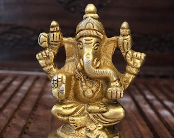 May include: A gold-colored metal statue of the Hindu deity Ganesha. The statue depicts Ganesha sitting with his trunk raised and his hands in a mudra. The statue is detailed with intricate carvings and is set on a round base.
