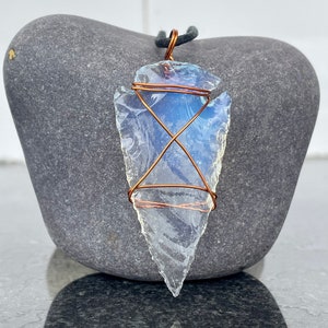 May include: A clear, arrowhead-shaped pendant with a copper wire wrap and a black cord. The pendant has a blue and white iridescent sheen. The arrowhead is resting on a grey stone.