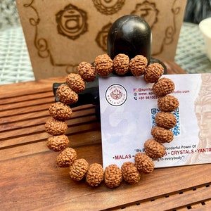 May include: A brown beaded bracelet made of textured, round beads. The bracelet is displayed on a wooden surface, with a black stone and a card with the text "SPIRITUAL WORLD STUDIO" in the background.