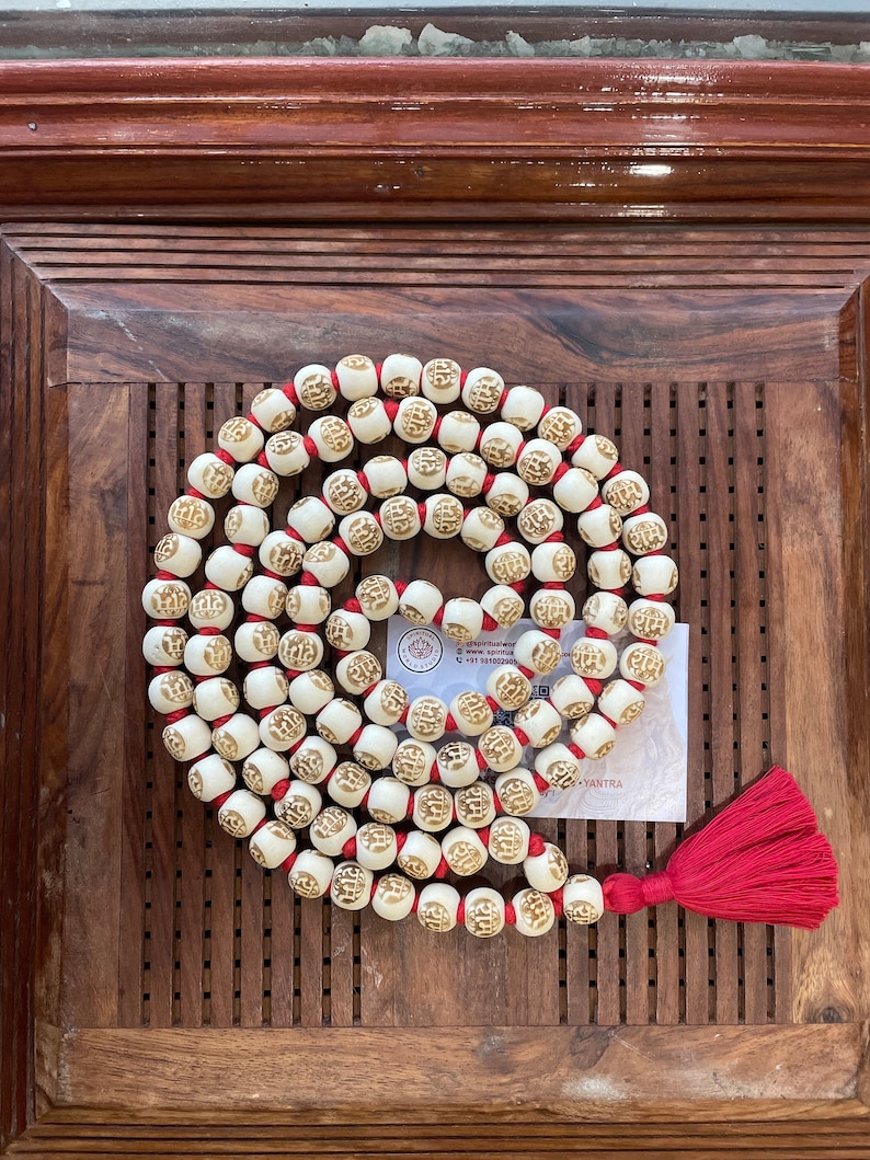May include: A white mala with red string and a red tassel. The beads are round and have a raised design. The mala is on a wooden surface.