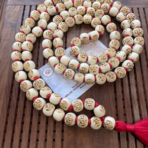 May include: A white mala necklace with red string and a tassel. The beads are carved with the word "राम" in Sanskrit. The necklace is on a wooden surface.