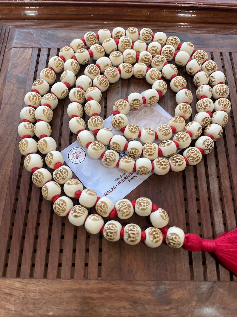 May include: A white mala necklace with red string and a tassel. The beads are carved with the word "राम" in Sanskrit. The necklace is on a wooden surface.