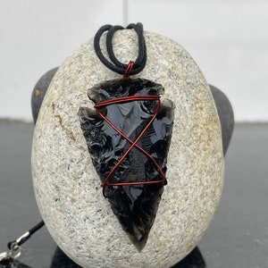 Puede incluir: Un colgante de punta de flecha de obsidiana negra envuelto en alambre de cobre, colgando de un cordón negro.