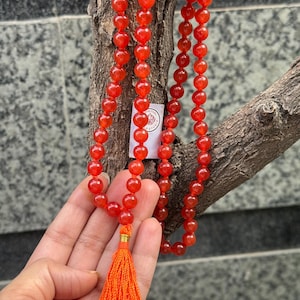 May include: A long strand of red carnelian beads with a tassel at the end. The beads are strung on a cord and are all the same size. The tassel is orange and is made of a soft material.