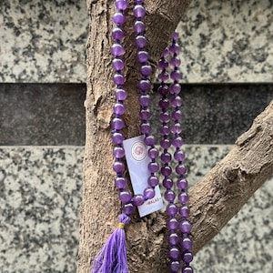 Può includere: Un mala in ametista viola con un fiocco, appeso a un ramo d'albero. Il mala ha 108 perle ed è infilato su un cordoncino viola. L'etichetta sul mala recita "MALAS."