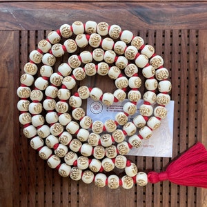 May include: A white mala with red string and a tassel. The beads are carved with the word "राम" in gold lettering. The mala is on a wooden surface.