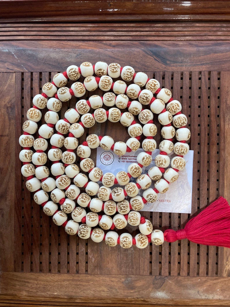 May include: A white mala with red string and a tassel. The beads are carved with the word "राम" in gold lettering. The mala is on a wooden surface.