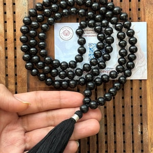 Puede incluir: Un collar de mala con cuentas negras y una borla negra. Las cuentas redondas parecen estar hechas de un material pulido. La borla est&aacute; unida con una pieza de metal plateado. Una tarjeta de visita est&aacute; en el fondo.