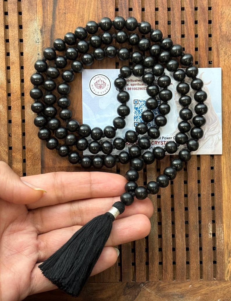 Puede incluir: Un collar de mala con cuentas negras y una borla negra. Las cuentas redondas parecen estar hechas de un material pulido. La borla est&aacute; unida con una pieza de metal plateado. Una tarjeta de visita est&aacute; en el fondo.