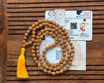 May include: A brown wooden mala with a yellow tassel. The mala is made of 108 beads and is used for meditation and prayer. The mala is on a wooden surface with a white card that says "ISO 9001:2015" and "Small & Medium Enterprises, Government of India".