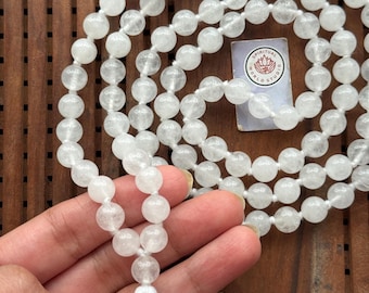 May include: A white beaded mala necklace with a white tassel and gold accents. The necklace is made of round, translucent beads. A small card with the text "SPIRITUAL WORLD STUDIO" is visible.