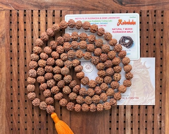 May include: A natural 7 Mukhi Rudraksha Mala, a strand of brown beads, with an orange tassel. The beads are arranged in a circular pattern on a wooden surface. The image includes a certificate from the Institute of Rudraksha & Gems Laboratory.