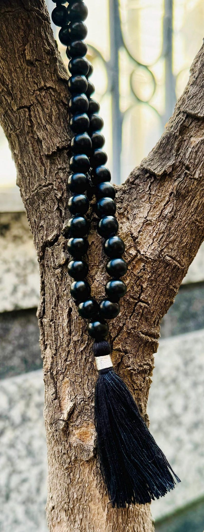 Puede incluir: Un collar de cuentas negras con una borla, colgado contra un tronco de &aacute;rbol texturizado. Las cuentas son redondas y espaciadas uniformemente. La borla est&aacute; hecha de hilos negros y est&aacute; unida al collar con una banda plateada.