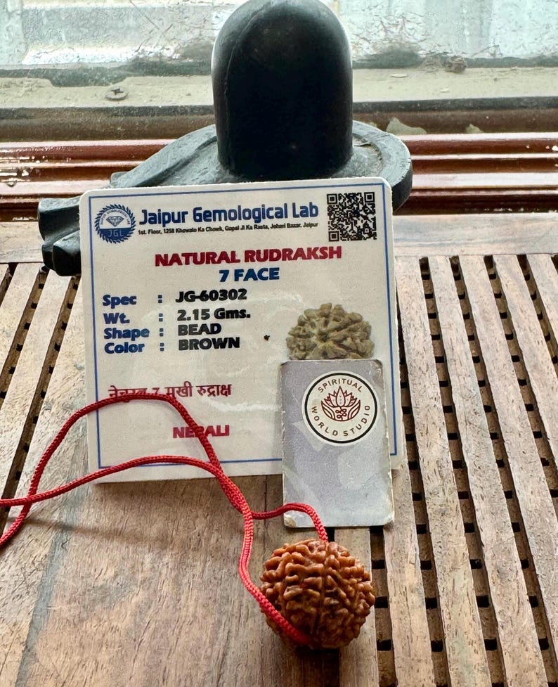 May include: A brown, natural Rudraksh bead necklace with a red cord. The image includes a card from Jaipur Gemological Lab with details: 7 Face, 2.15 grams, and brown color. A Spiritual World Studio card is also present.