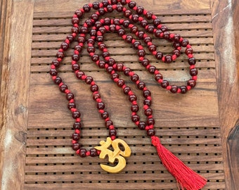 May include: A red wooden mala bead necklace with a wooden Om pendant and a red tassel. The necklace is on a wooden surface.