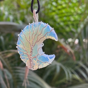 May include: A silver wire tree of life design is wrapped around a crescent moon shaped opal pendant. The pendant is hanging from a black cord.