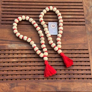 May include: Two wooden bead garlands with red string accents and red tassels. The garlands are on a wooden surface.